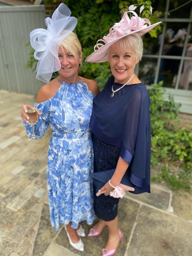 Two elegantly dressed women at a wedding, wearing stylish Mother of the Bride dresses. One is in a floral print dress and the other in a navy evening gown